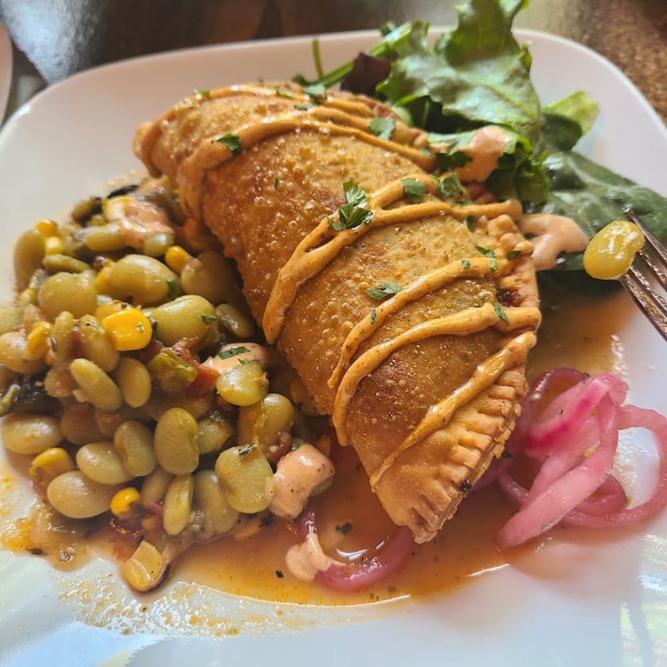 A golden-brown empanada drizzled with sauce, served with lima beans, corn, and pickled red onions.