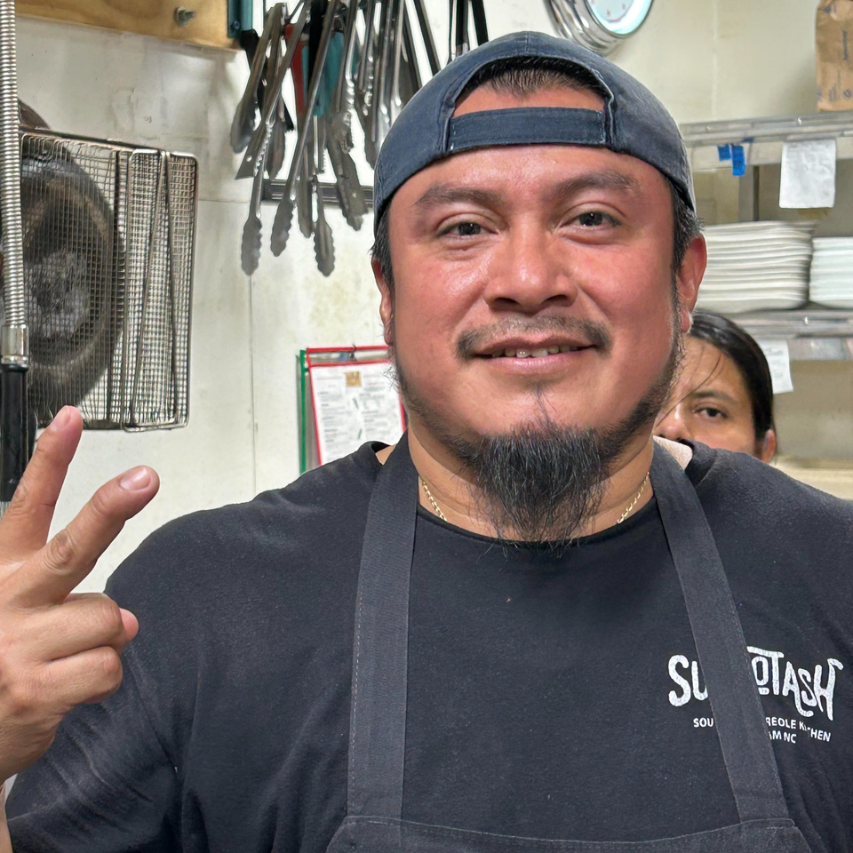 A smiling Succotash chef in a black apron and cap gives a peace sign in the kitchen.