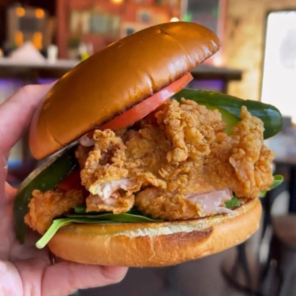 Hand holding a crispy fried chicken sandwich with lettuce, tomato, and pickles on a brioche bun.